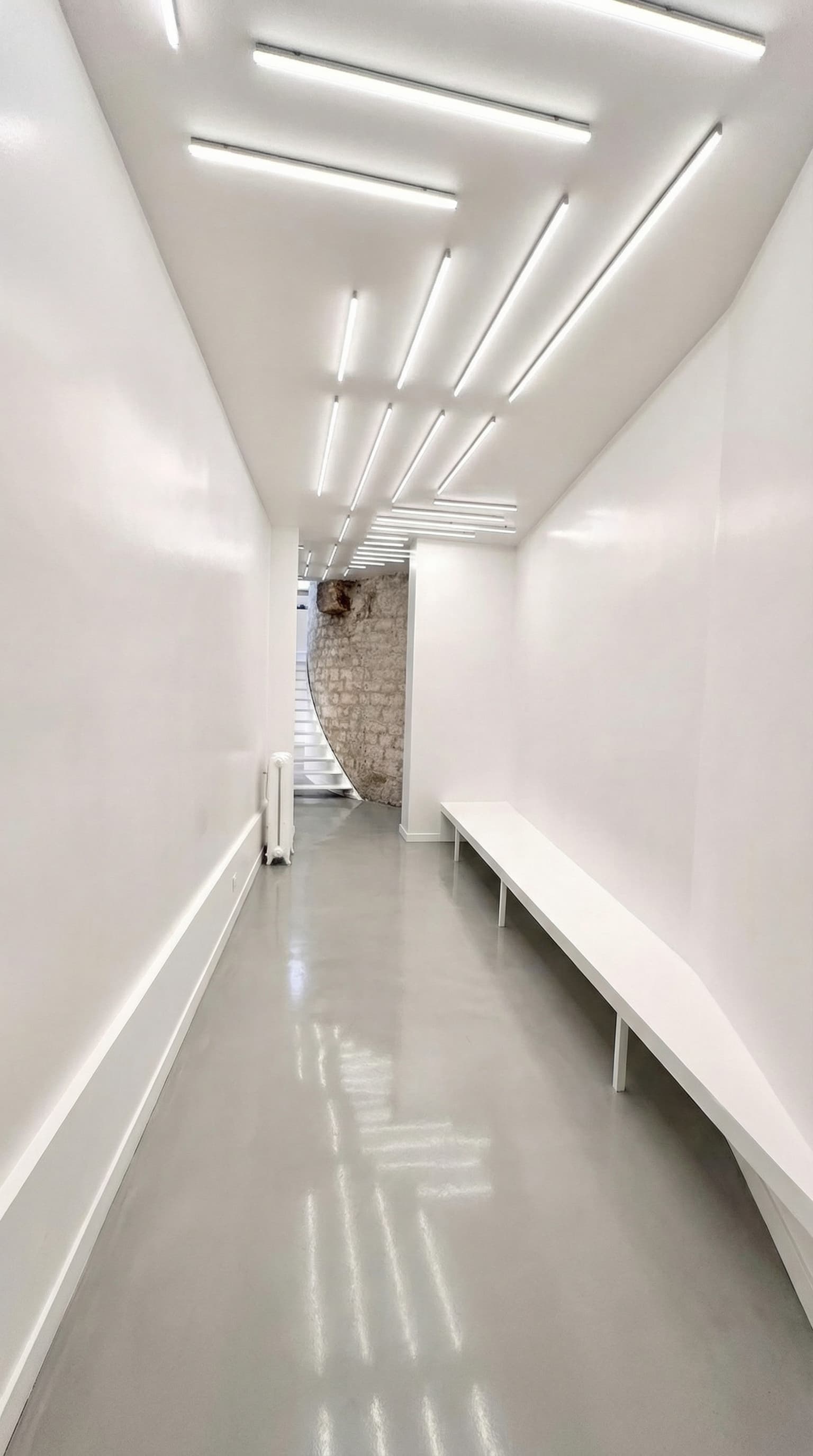 Corridor blanc avec néons linéaires et mur en pierre apparente